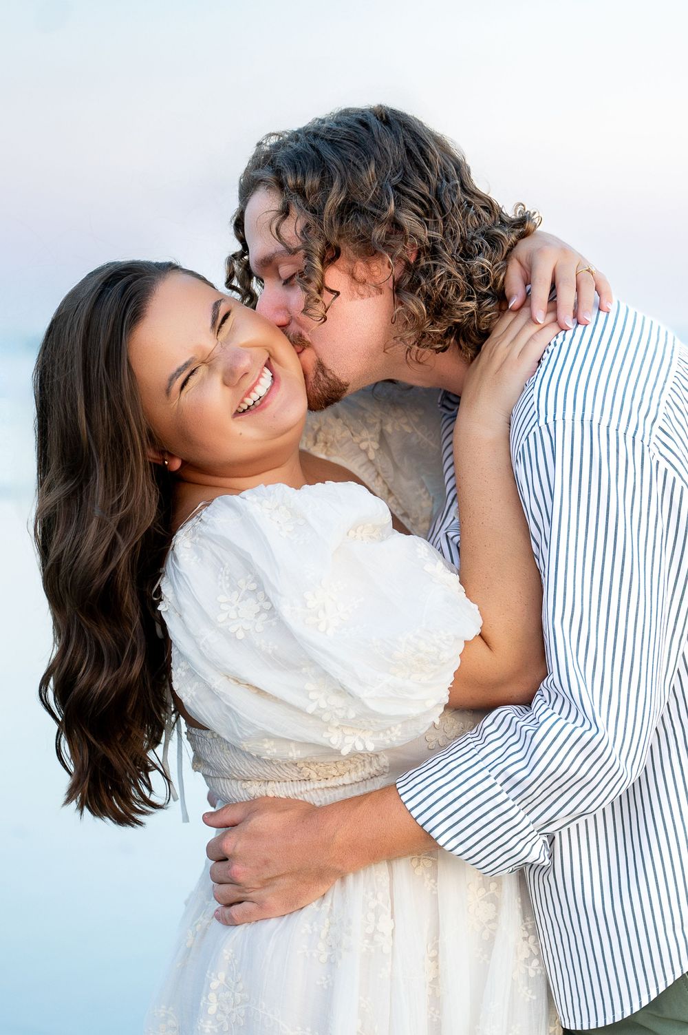 A romantic couple shares an intimate moment on the beach, with the woman in white and man in stripes.