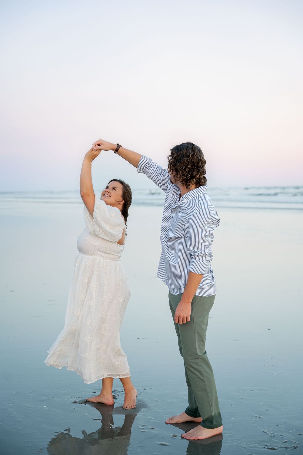 Two people dance joyfully on wet sand at the beach during golden hour.