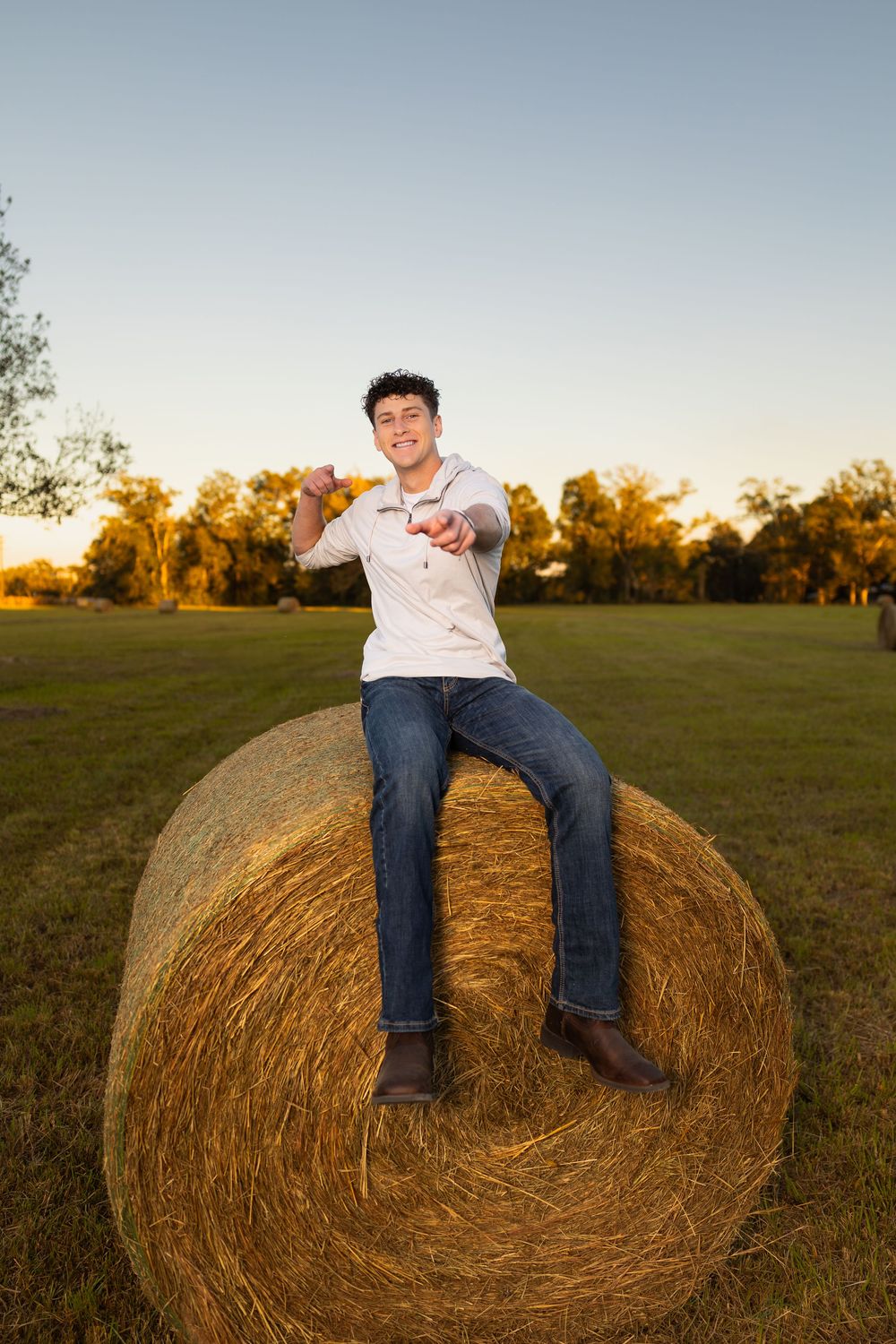 A person sits atop a round hay bale in a field during golden hour.