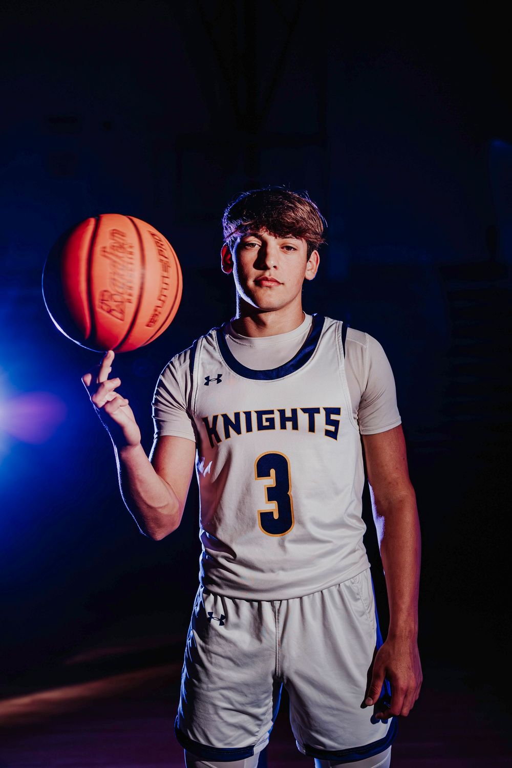Basketball player in white Knights jersey poses with ball in dramatic blue lighting.