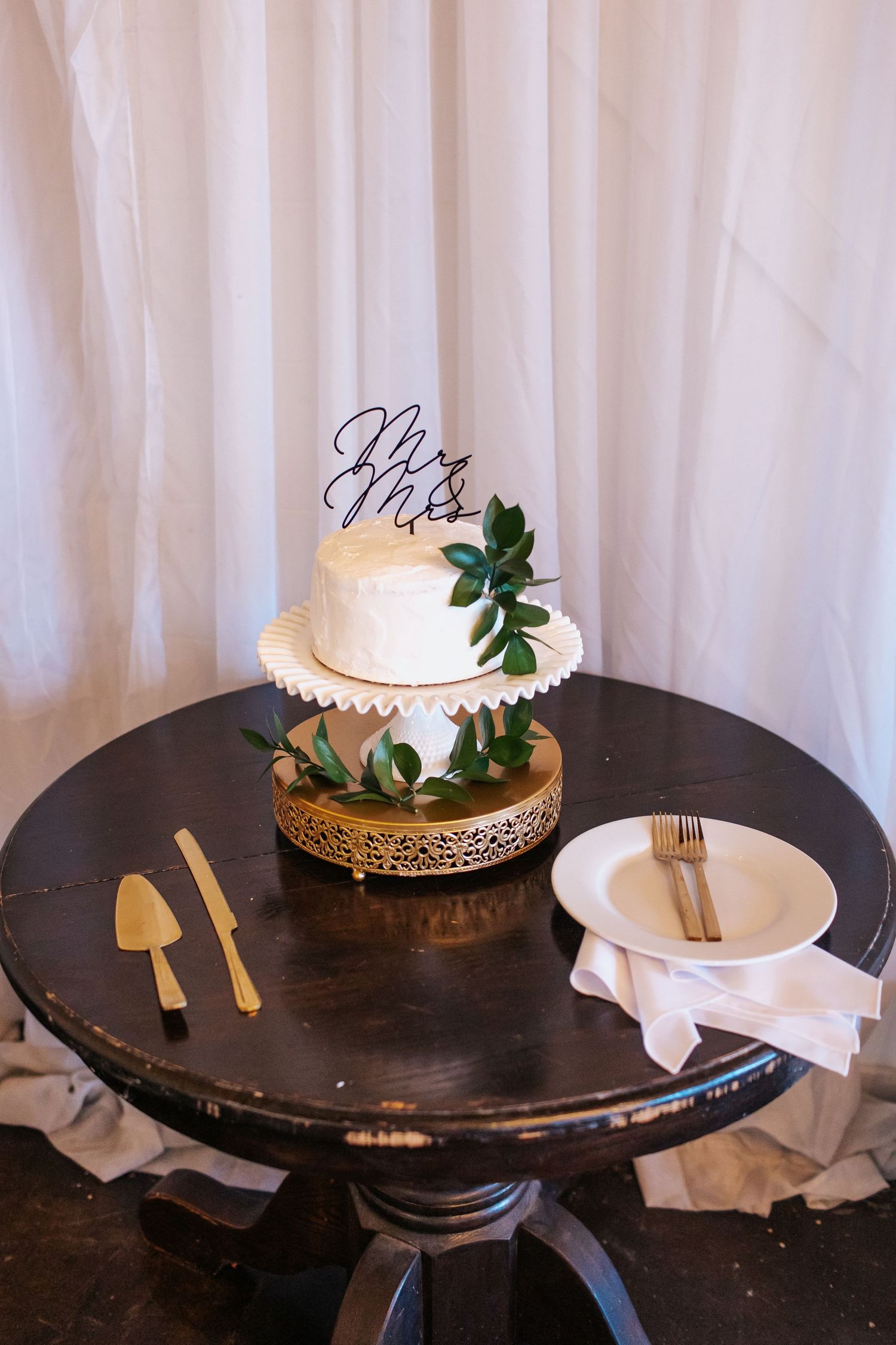 Simple white wedding cake decorated with greenery and Mr & Mrs topper on dark wooden table.