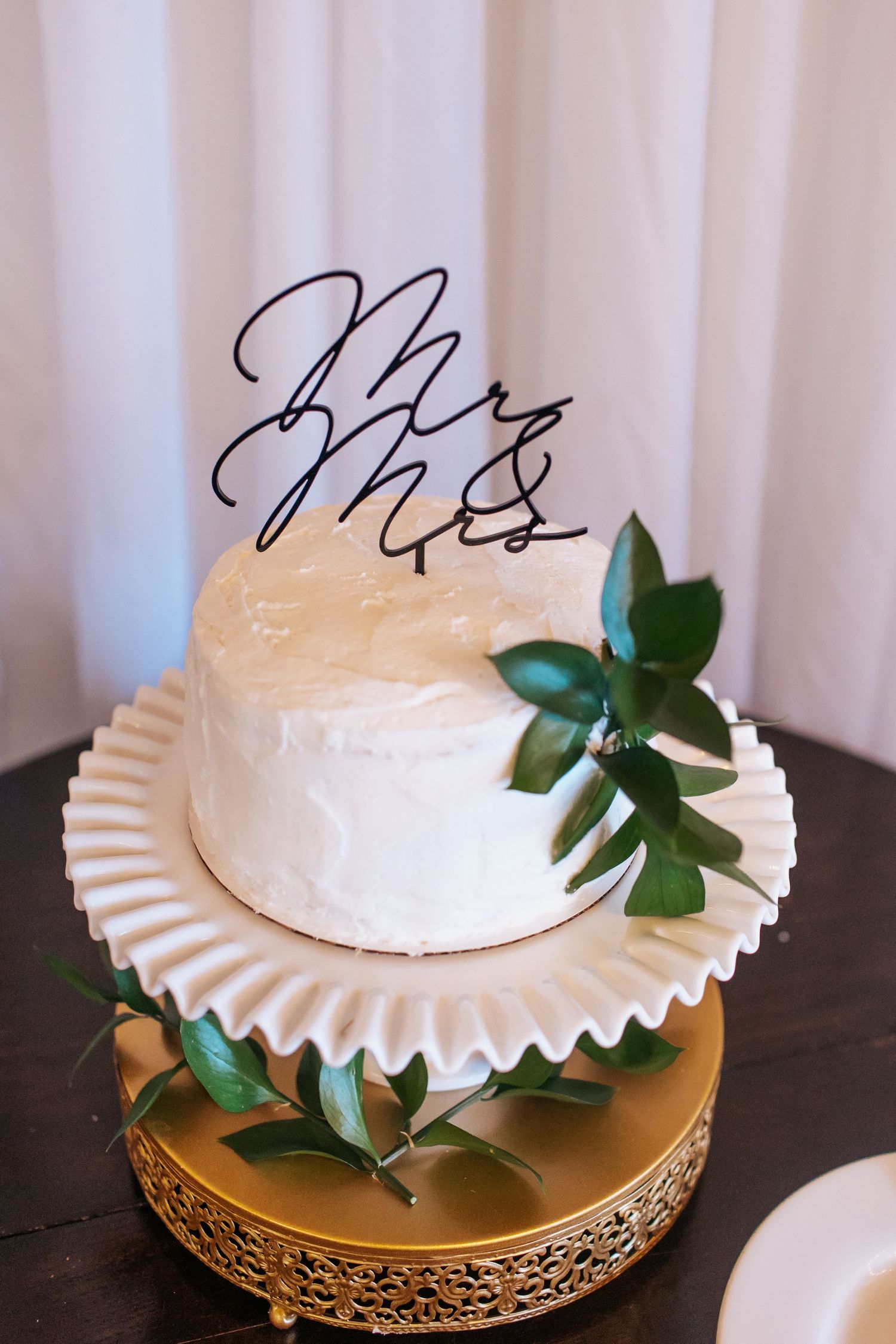Elegant white wedding cake with modern calligraphy topper and fresh green leaves.