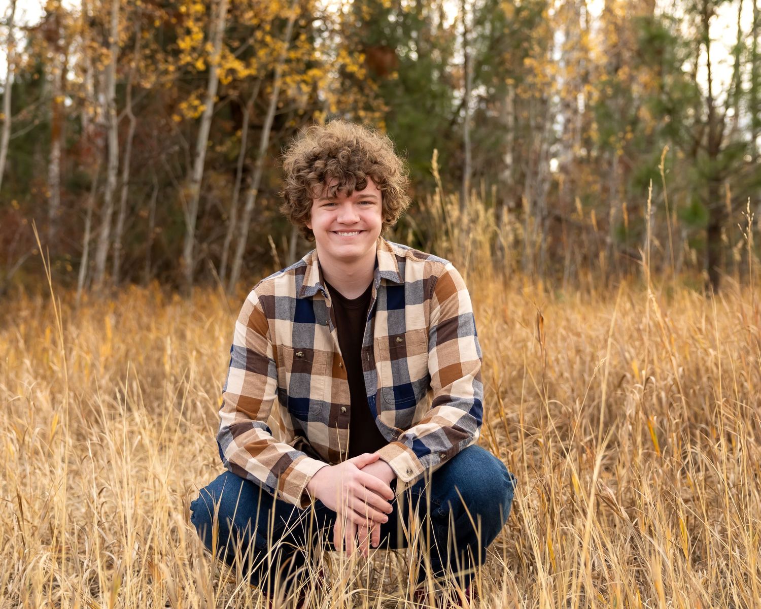 Someone in a plaid flannel shirt crouches in tall golden grass with autumn trees in the background.