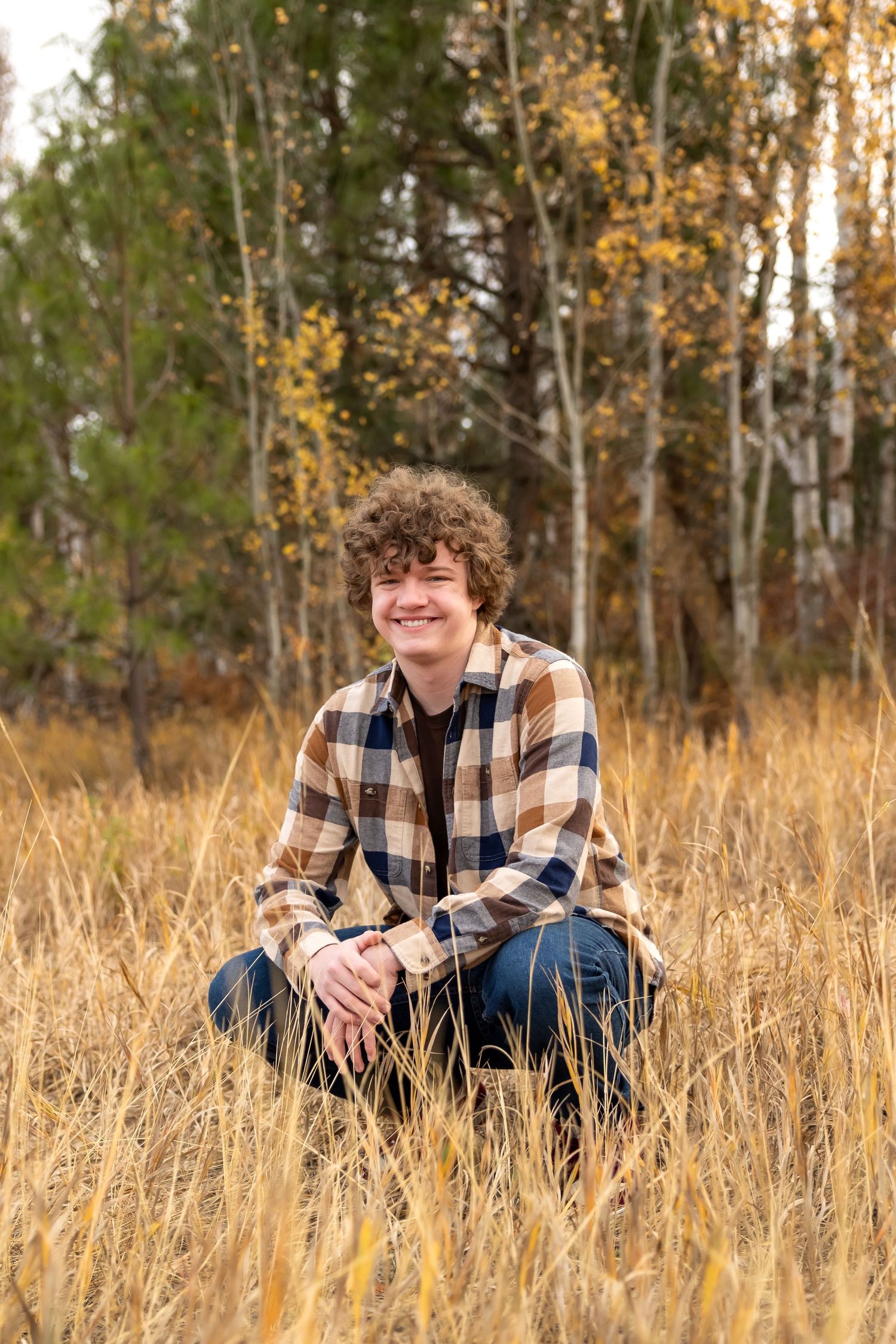 A person wearing a plaid shirt sits in golden autumn grass with trees in the background during sunset.