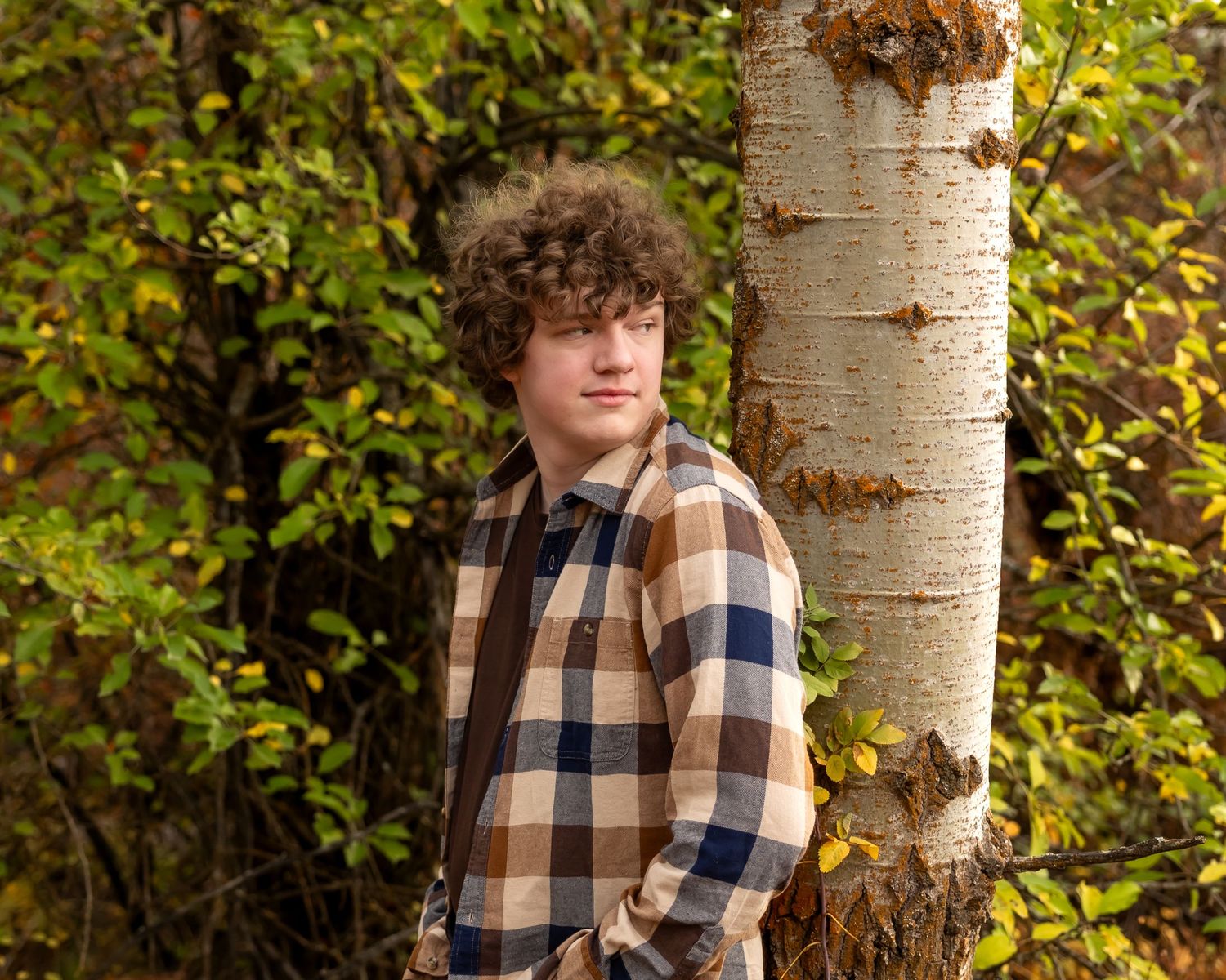 A person in a plaid flannel shirt poses against a white birch tree trunk with autumn foliage in the background.