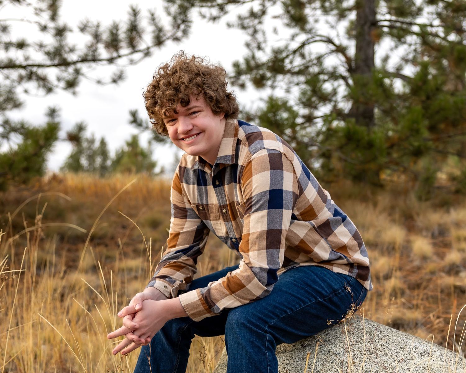 A plaid flannel shirt and jeans outfit photographed outdoors in golden grass with pine trees in the background.