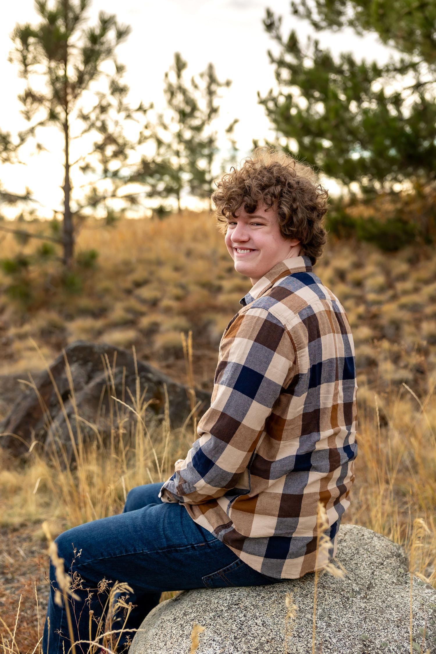 A person in a brown plaid flannel shirt and jeans sits on a large rock at sunset in a desert landscape.