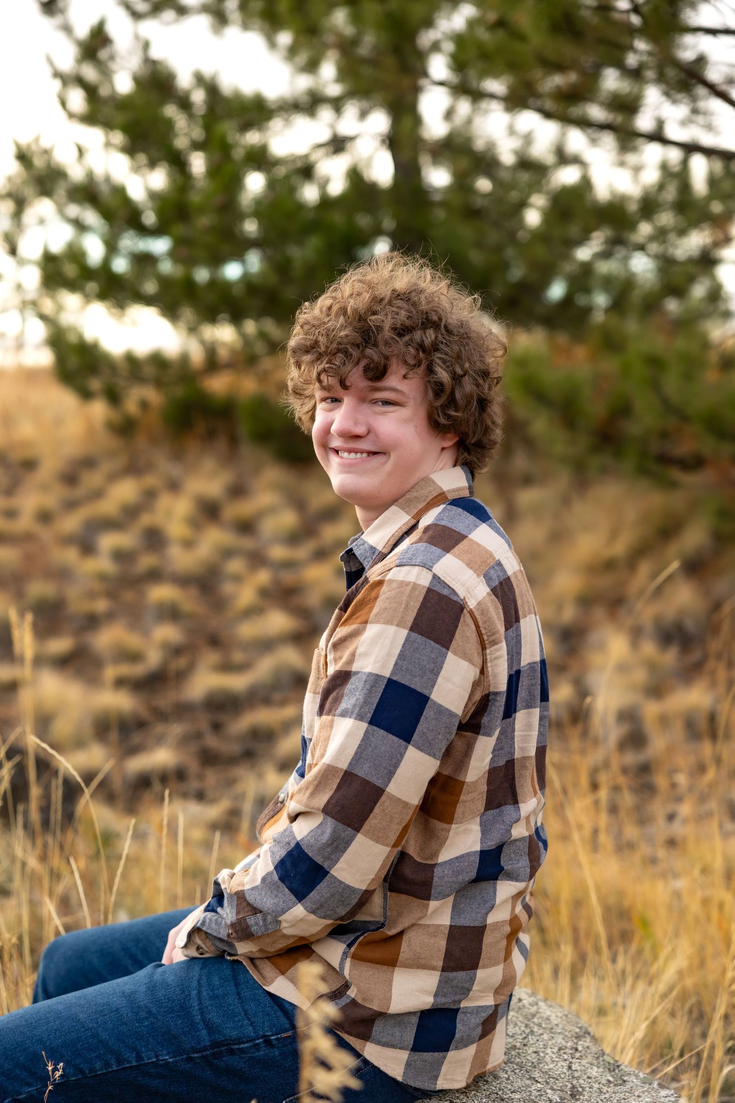 Portrait of someone in a plaid flannel shirt and jeans sitting on a rock during golden hour in nature.