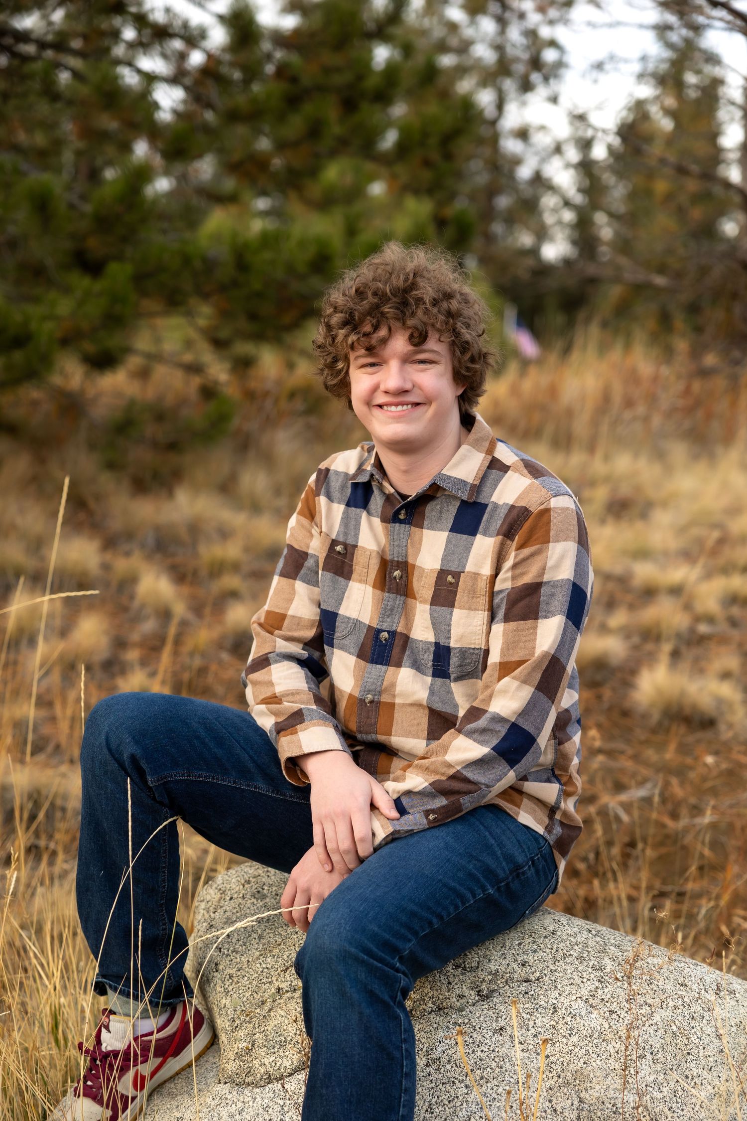 Person in plaid flannel shirt and jeans sits casually on rocky ground with autumn foliage in background.