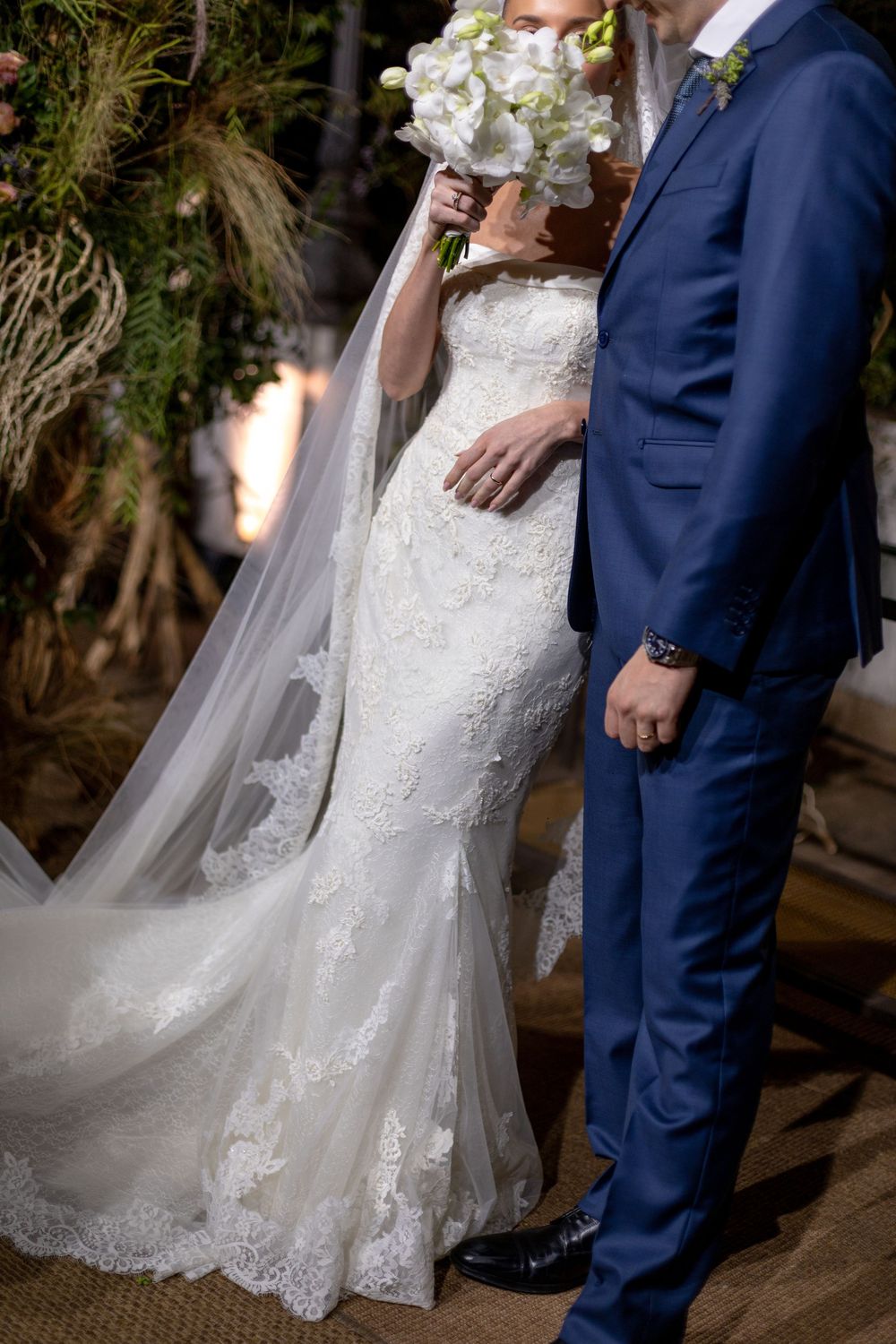 Close-up details of a bride in a white lace wedding gown standing next to a groom in a navy blue suit.