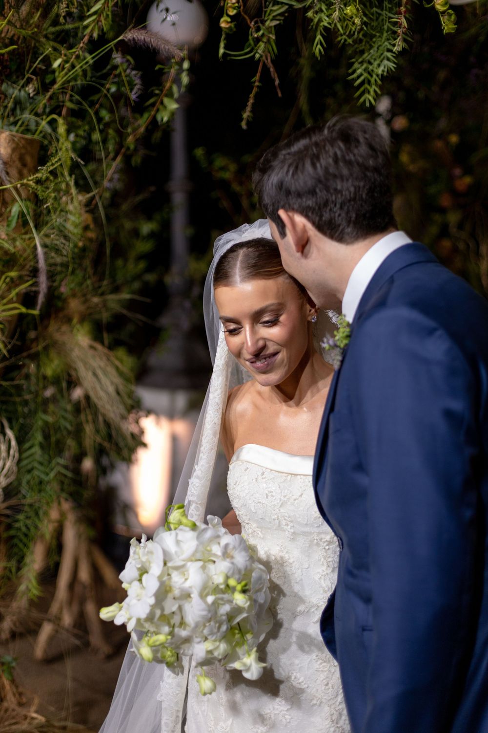 A couple shares an intimate moment during their wedding ceremony under lush green trees.