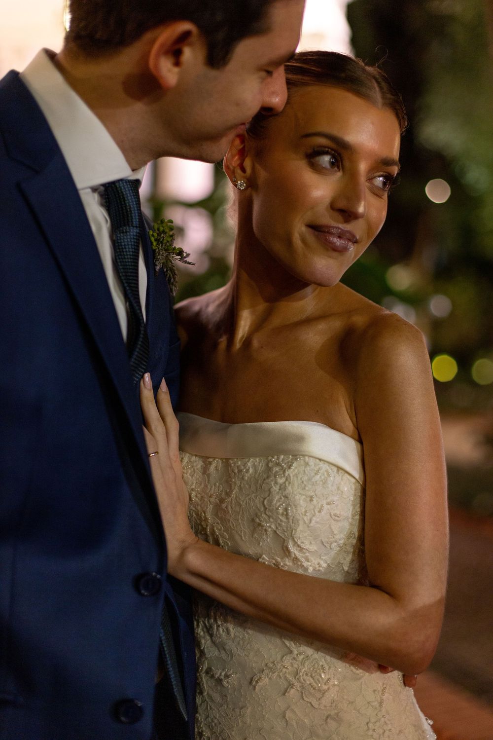 Romantic wedding couple embraces during outdoor evening reception with soft bokeh lights in background.