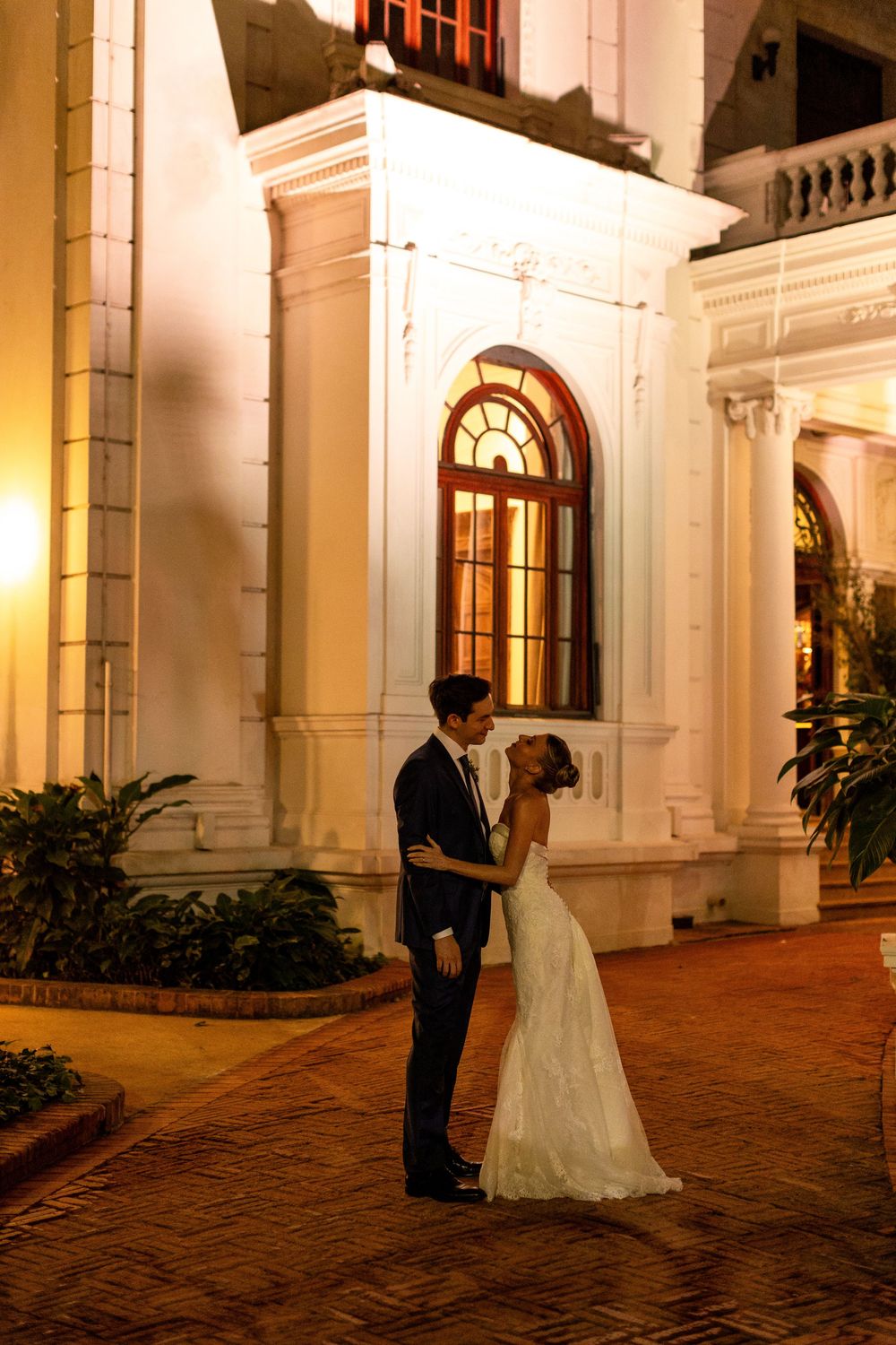 A romantic moment between newlyweds on a brick pathway outside an elegant colonial-style building at dusk.