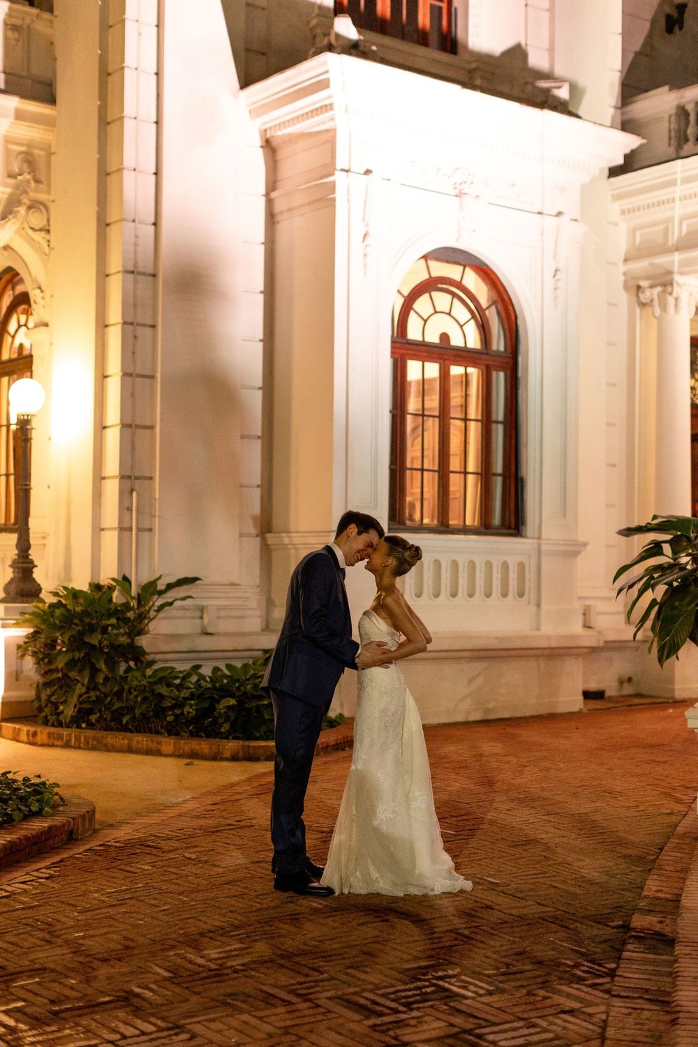 A romantic wedding couple shares an intimate moment on a brick courtyard in front of an elegant white colonial building at night.