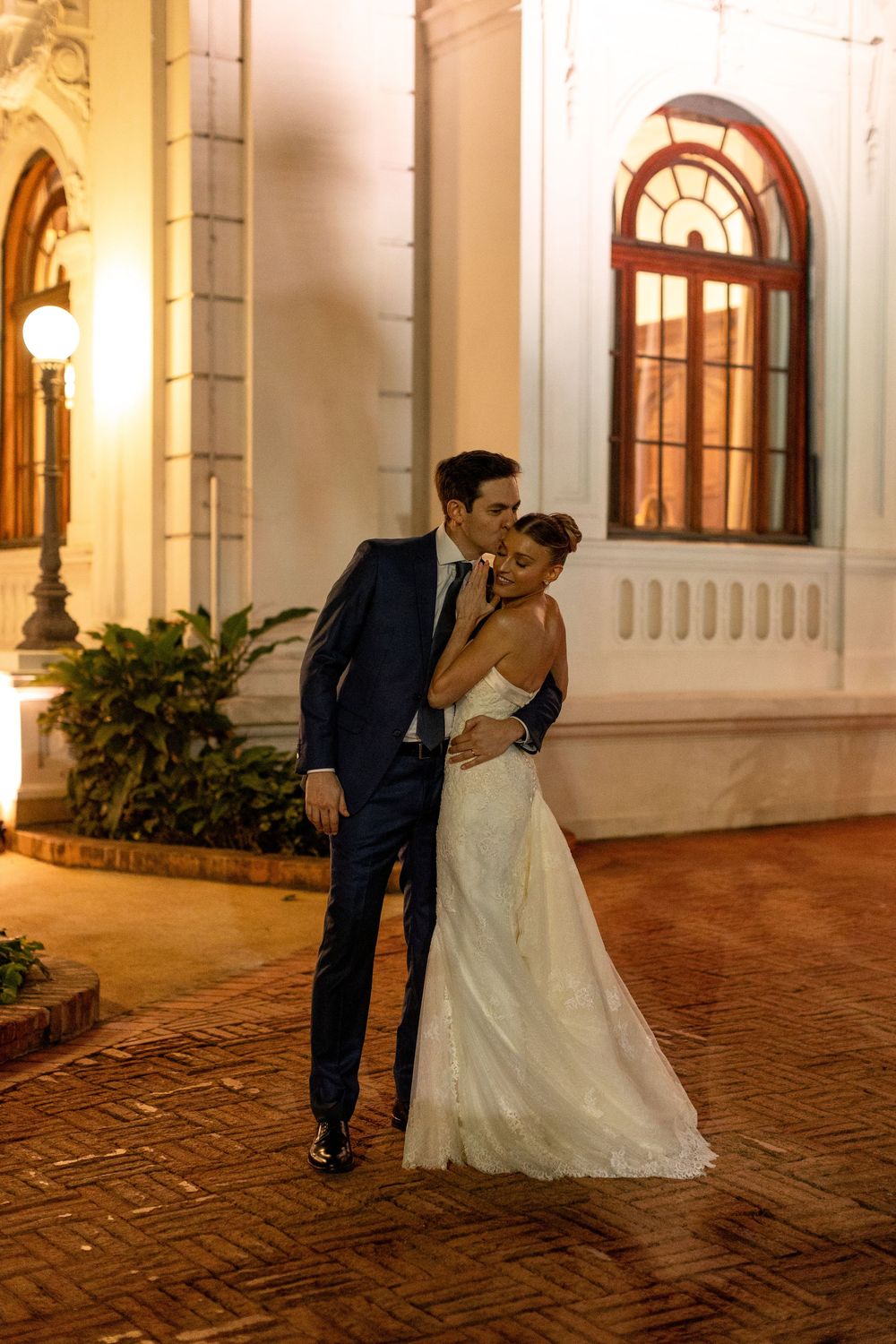 A couple shares an intimate moment in their wedding attire outside a grand building with arched windows and warm lighting.