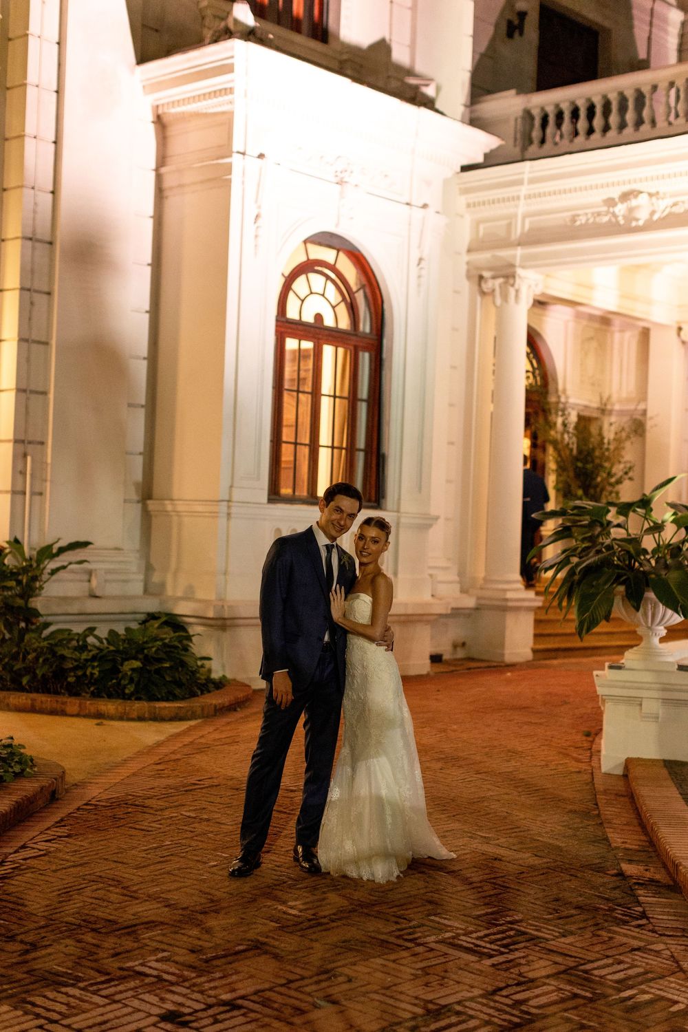 A newlywed couple stands close together on a brick patio against a beautifully lit colonial-style building backdrop.