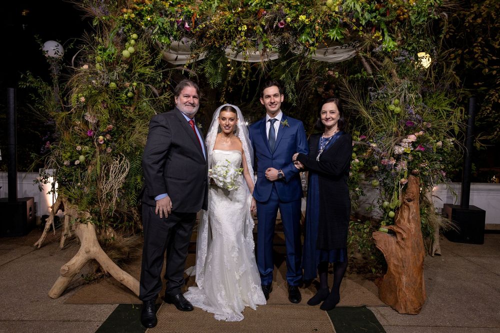 Wedding party of four people posing together under an archway decorated with greenery and lights.
