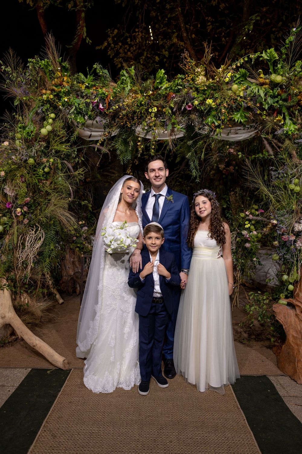 A wedding party poses under a lush floral archway decorated with greenery and flowers at an evening ceremony.