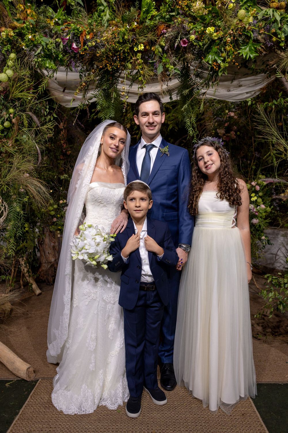 Wedding party poses together in garden setting with lush greenery backdrop and rustic stone wall.
