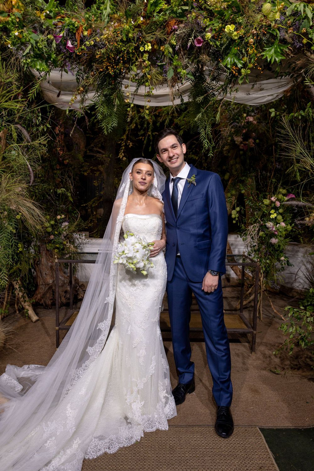 A couple in wedding attire poses in a garden setting with lush greenery and vines creating a romantic backdrop.