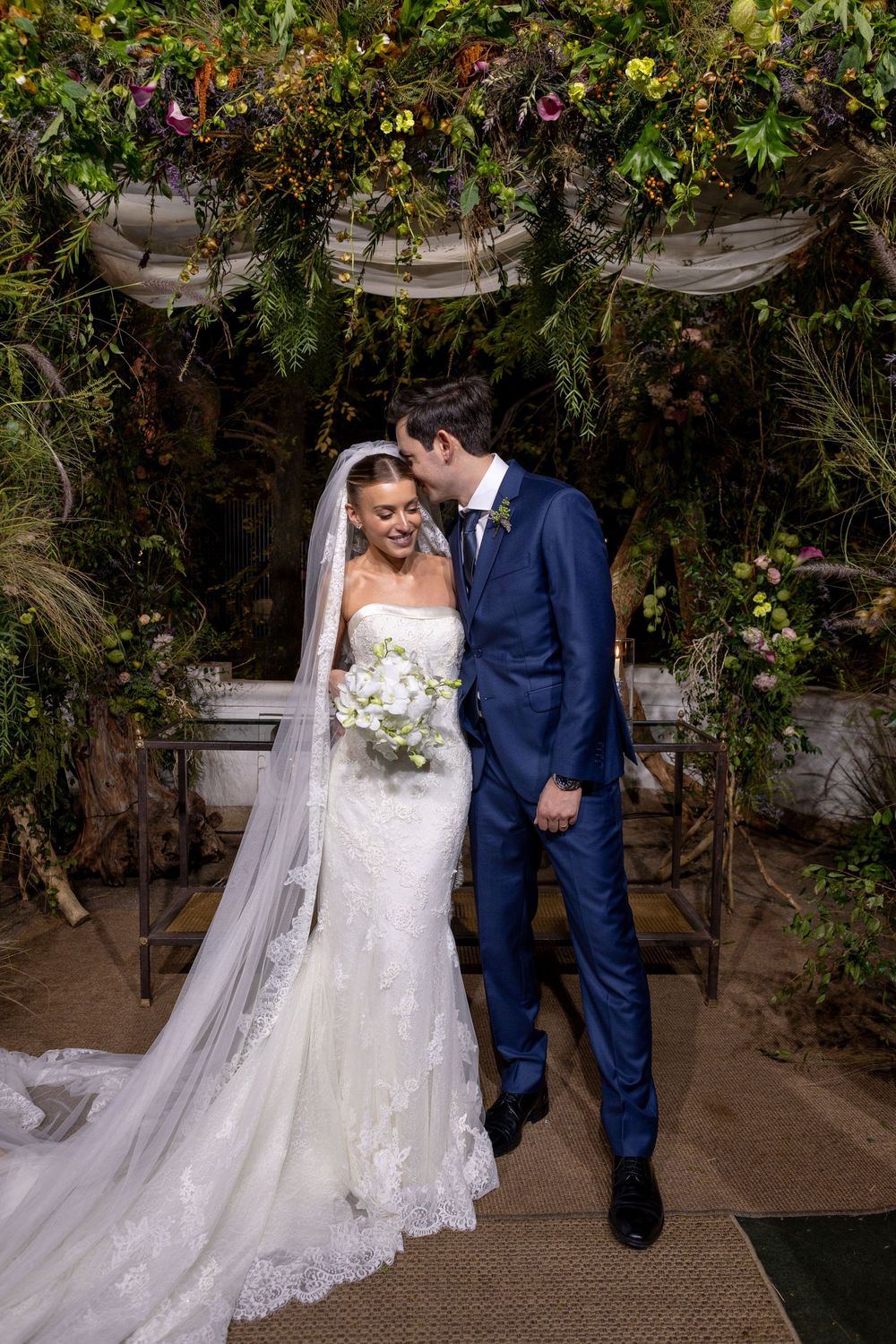 Bride in flowing white gown and groom in navy suit share tender moments under a lush garden archway.