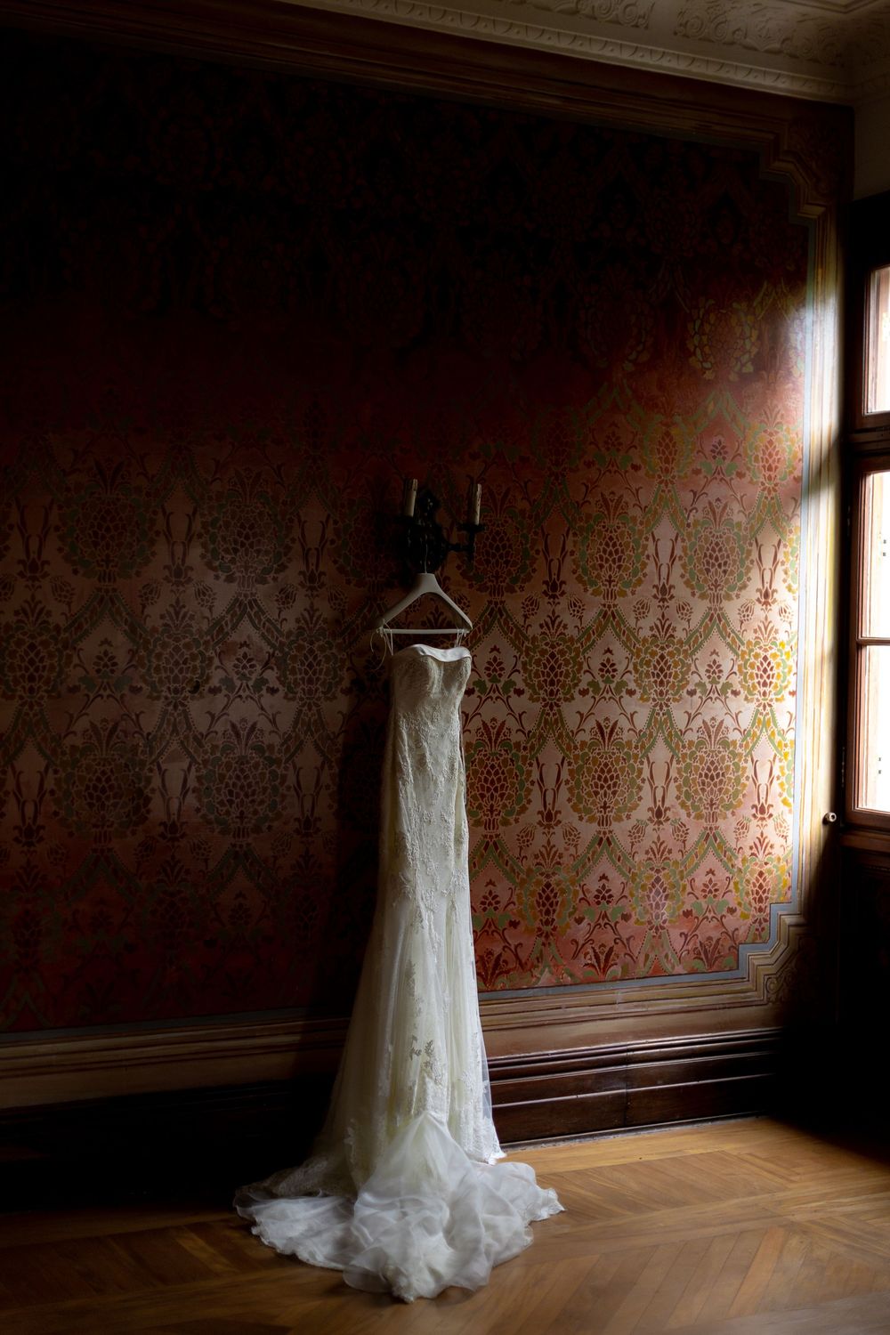 Elegant white wedding dress hanging against ornate red damask wallpaper in a vintage room.
