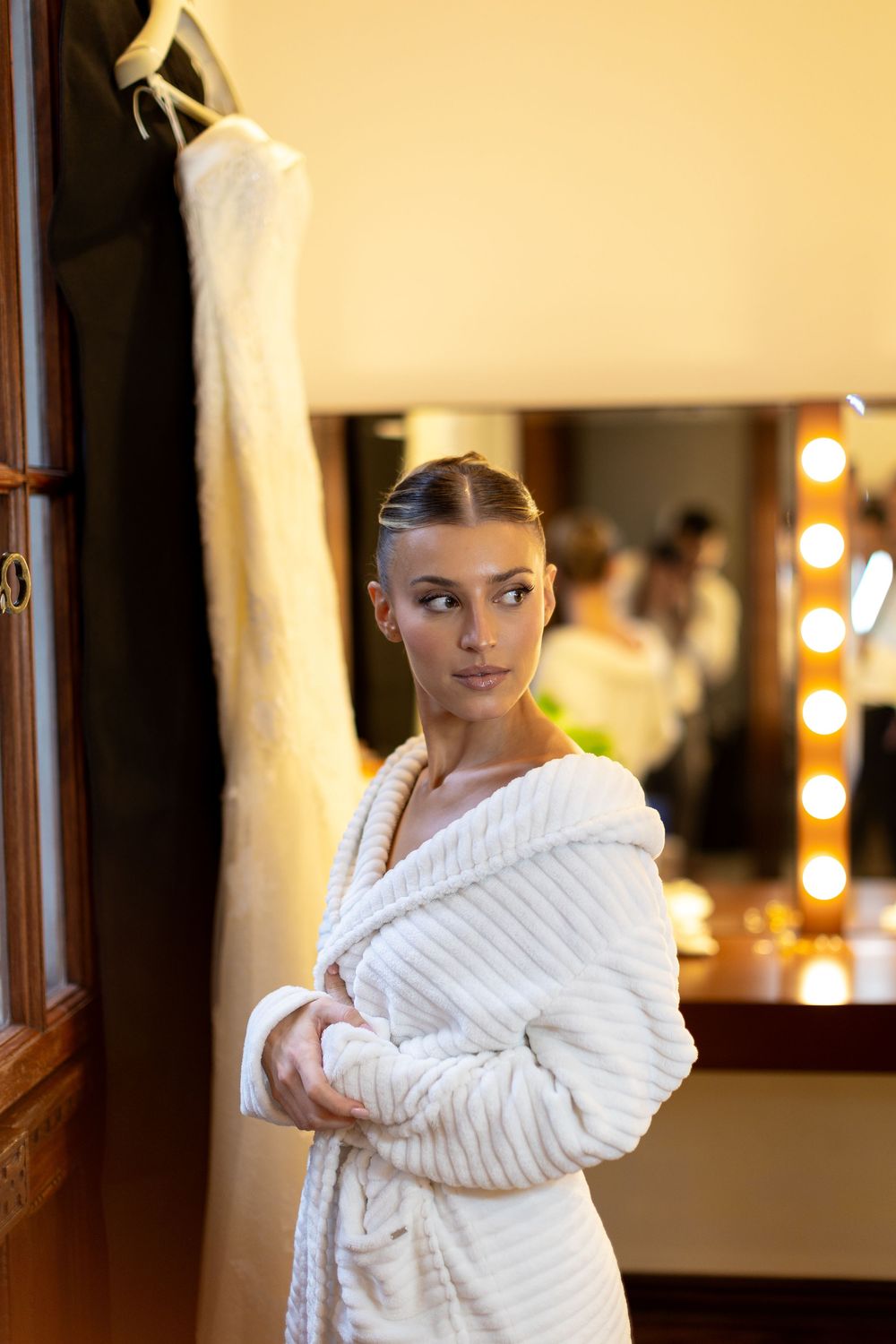 A person wearing a plush white bathrobe poses in front of a vanity mirror with glowing lights.