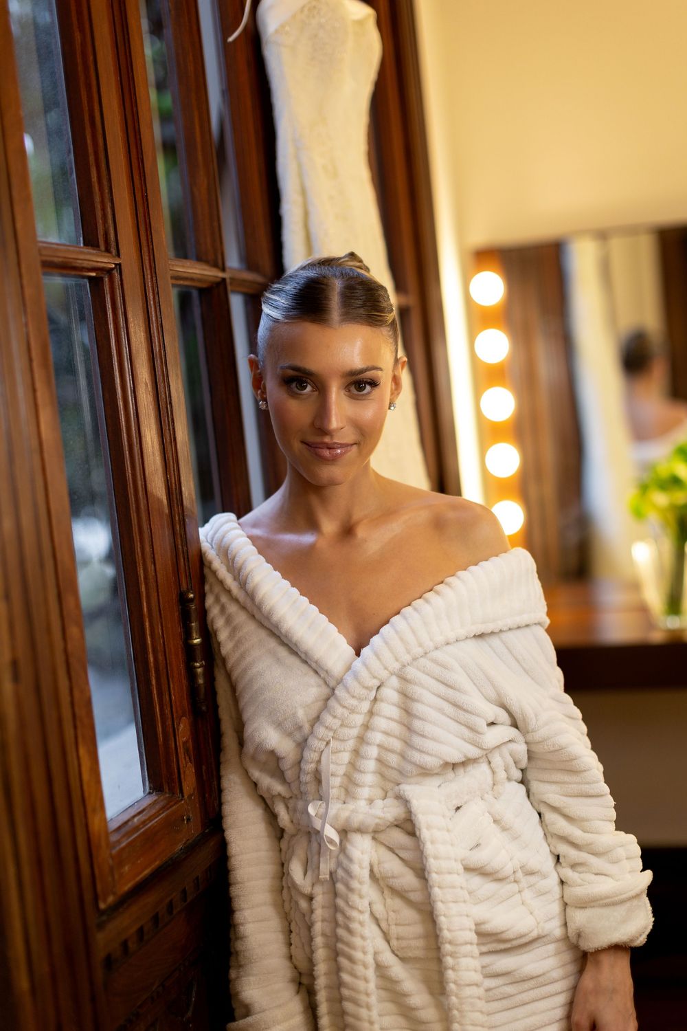 Bride in a textured white robe poses elegantly in a warmly lit dressing room.