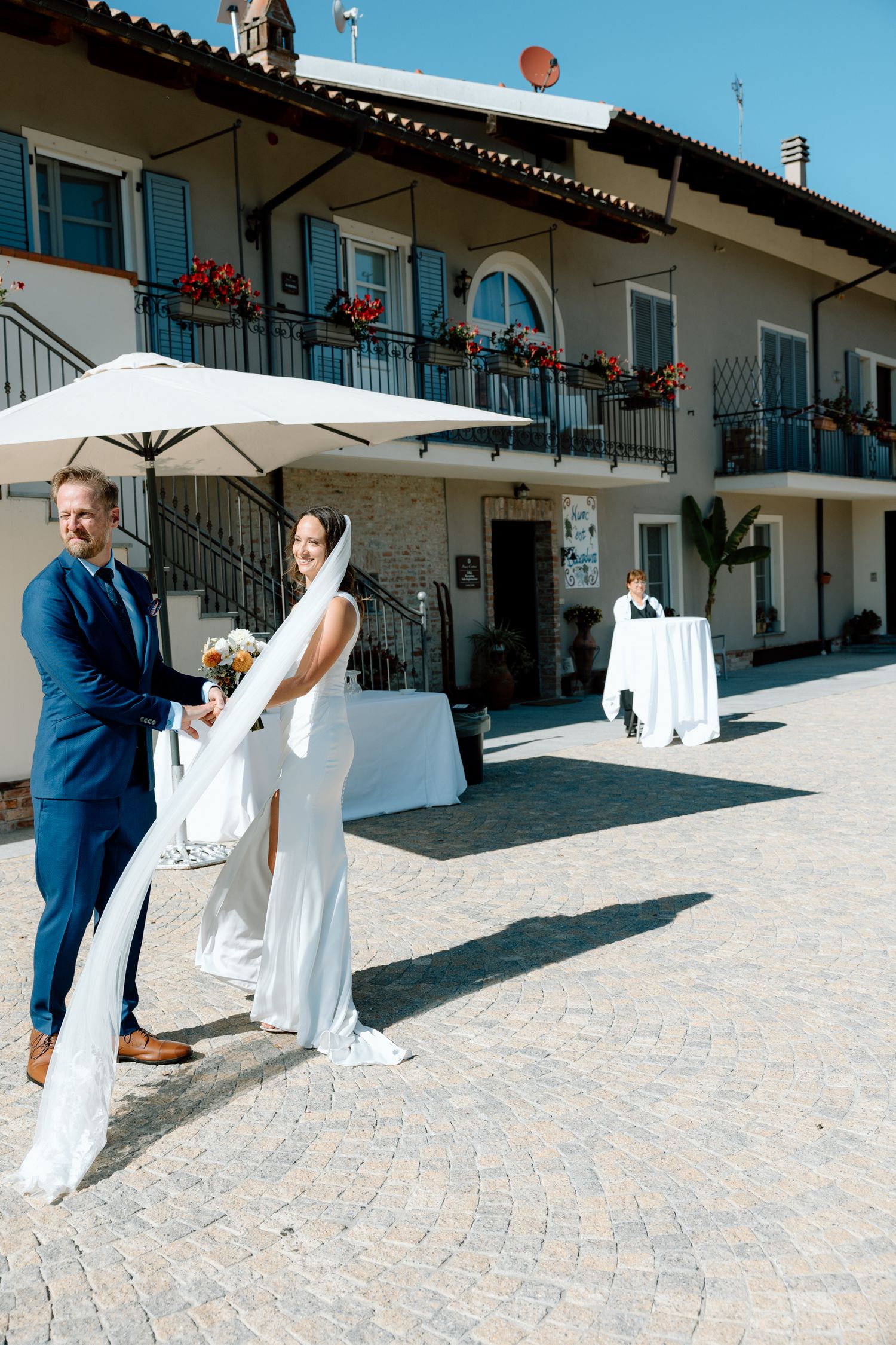 A couple in wedding attire share an intimate kiss in front of a building with blue shutters and white walls.