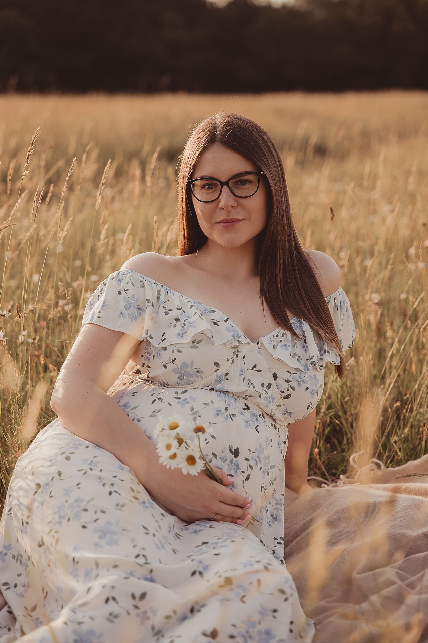 A pregnant woman in a white floral dress sits peacefully in a golden sunlit field of tall grass.