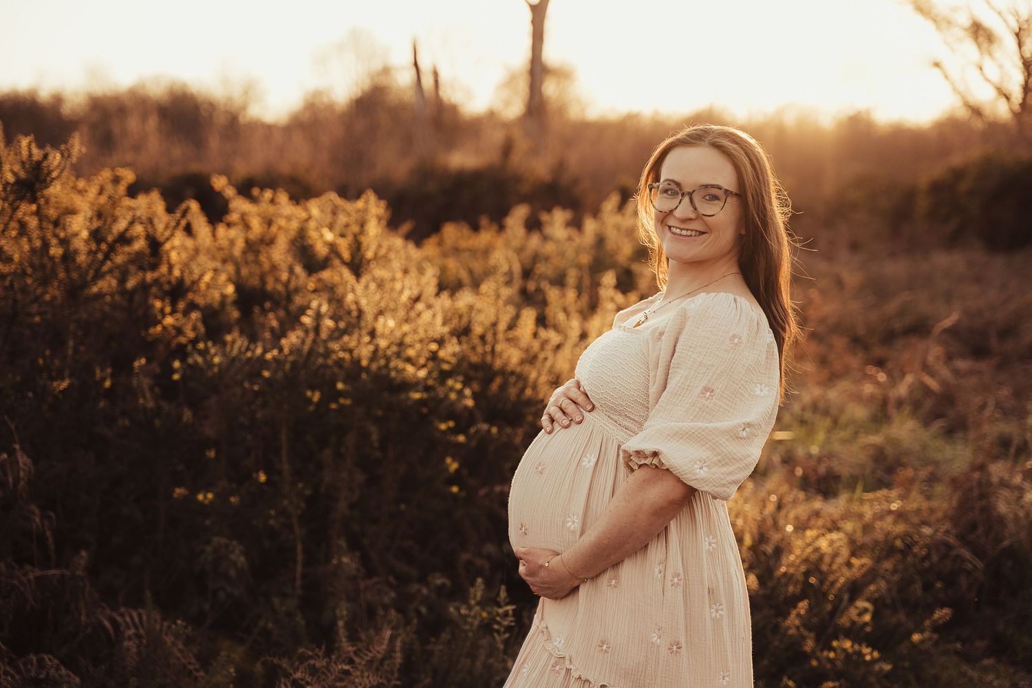 Pregnant woman in white dress holding baby bump at golden hour in a field.