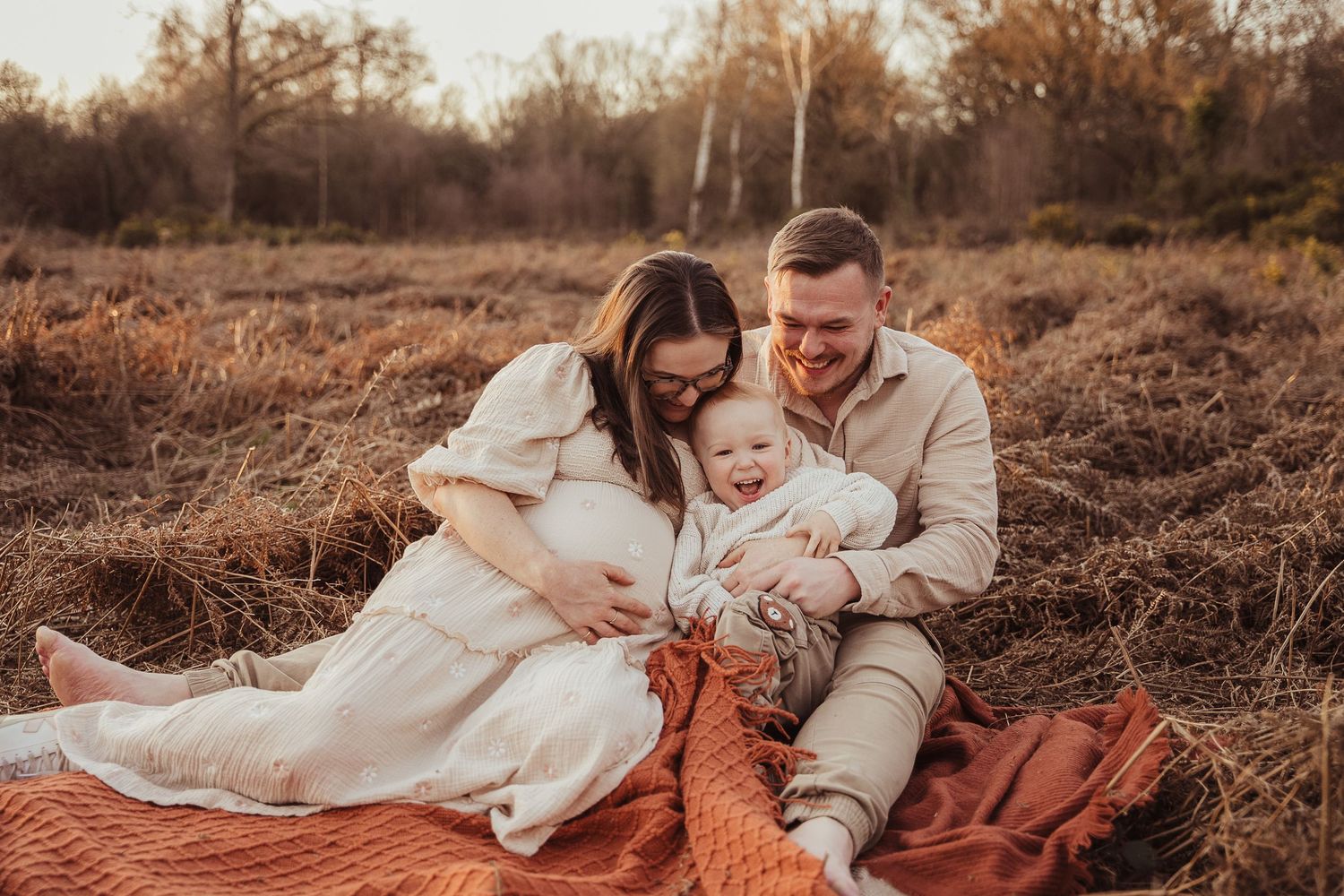 A happy family sits on a blanket in a golden field, parents smiling at their baby during an autumn outdoor photo session.