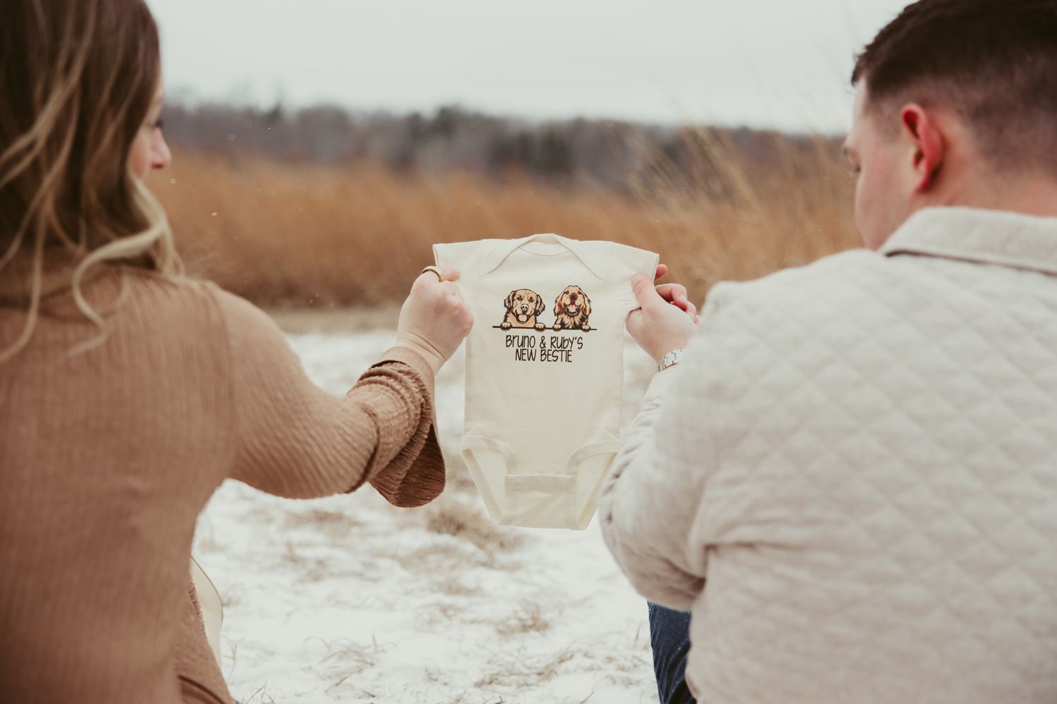 Couple holding up baby onesie with teddy bear design during winter pregnancy announcement.