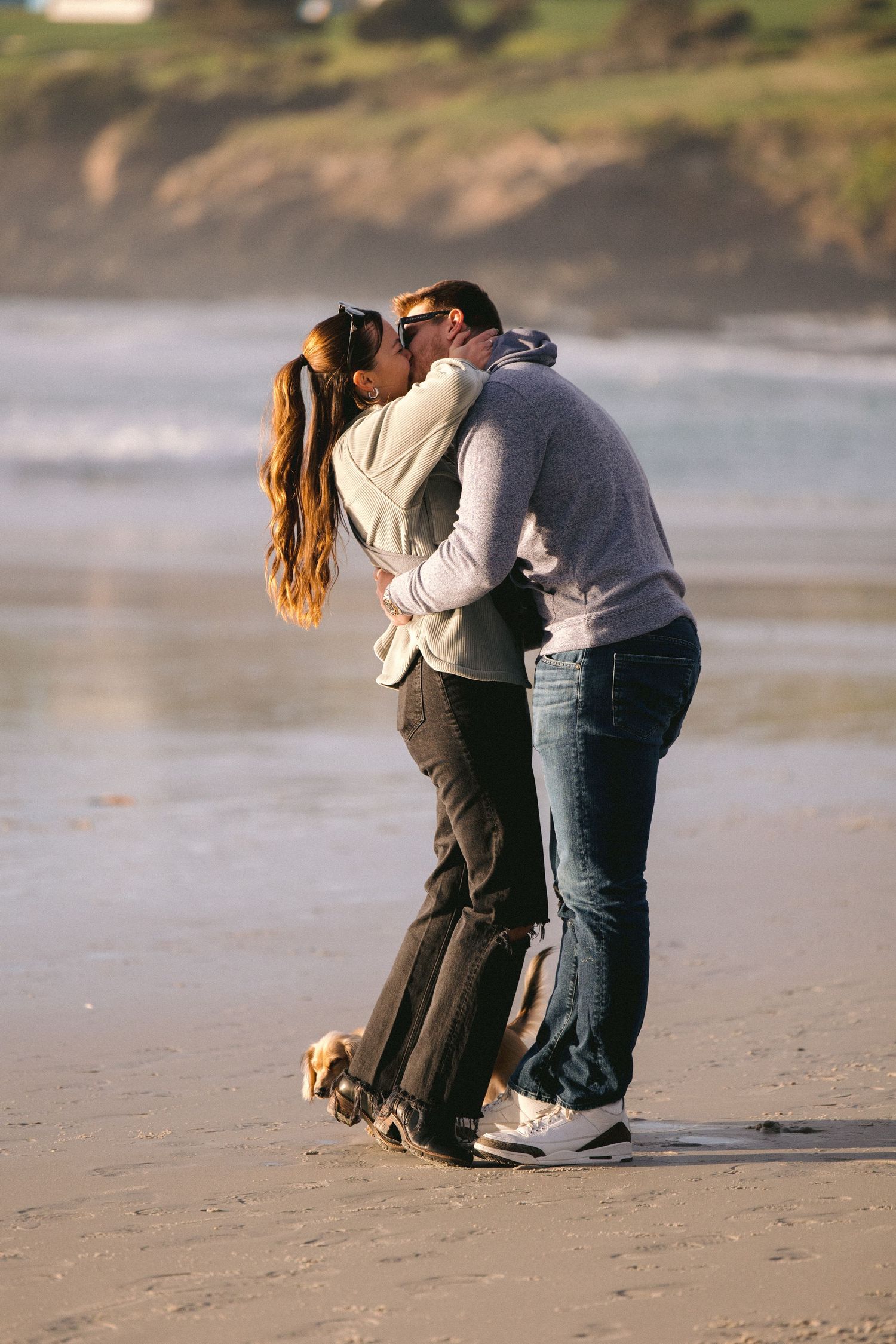 A couple shares a romantic kiss on a peaceful beach at sunset with waves and cliffs in the backdrop.