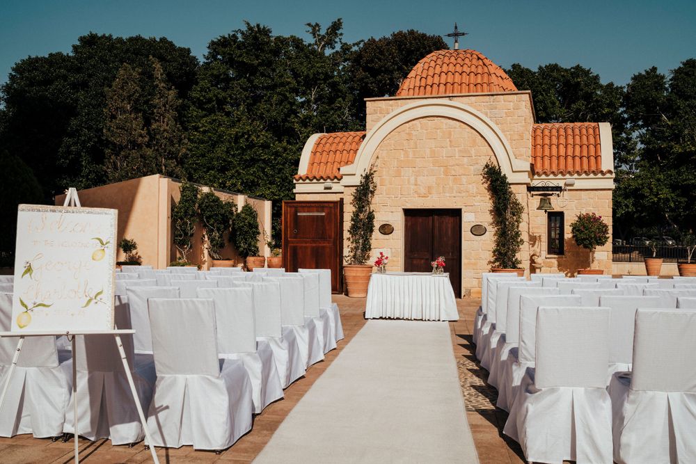 A Mediterranean-style wedding ceremony setup with white chairs and a terracotta-domed gazebo under blue skies.