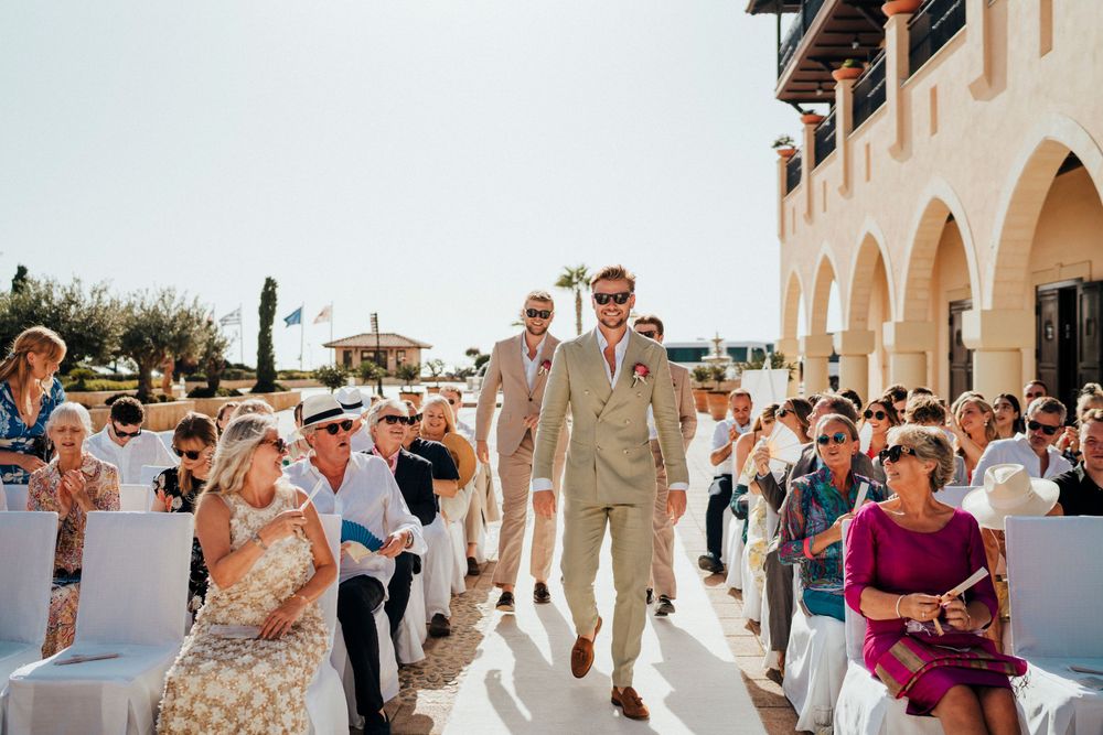 Wedding guests walk along a sun-drenched courtyard with Mediterranean-style arches during an outdoor celebration.
