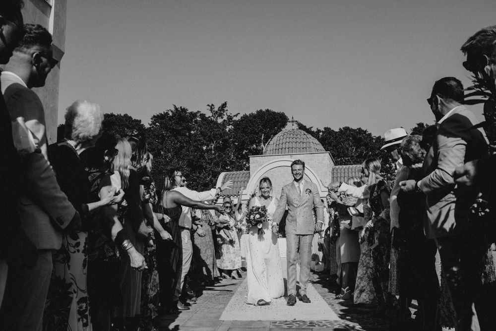 Wedding guests line a pathway and throw petals as newlyweds walk between them in black and white photograph.