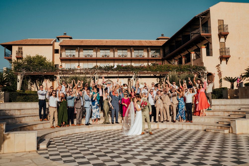 Wedding guests gather on a checkered outdoor patio in front of a Mediterranean-style venue during sunset.