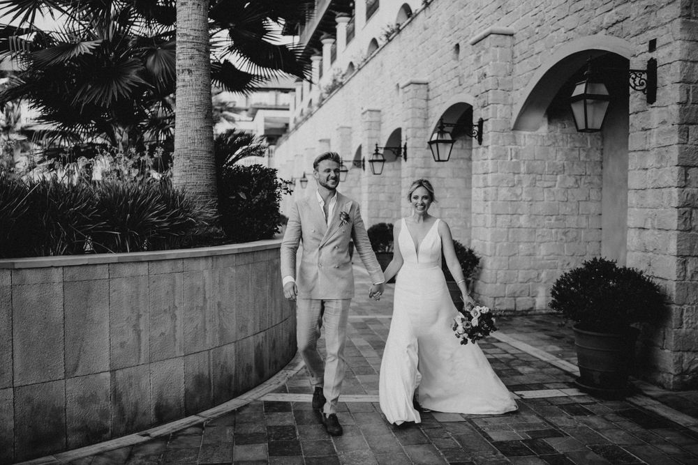 A black and white wedding photograph of a couple walking down a palm-lined path beside a Mediterranean-style building.
