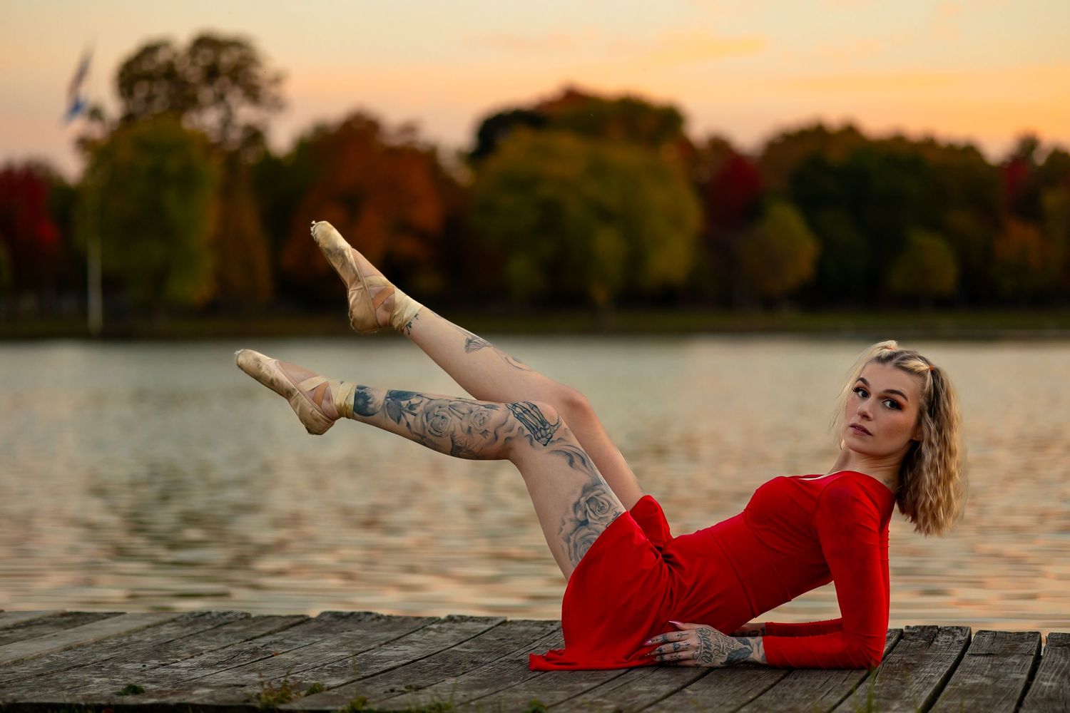 Dancer in red dress strikes elegant pose on wooden dock during sunset.