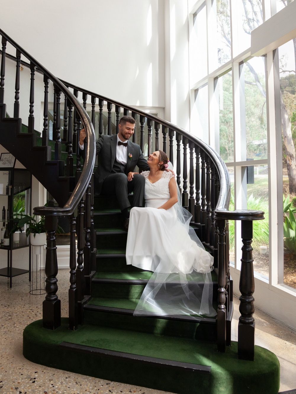 Bride and groom sitting together on an elegant curved staircase with dark railings and green carpet in a bright venue.