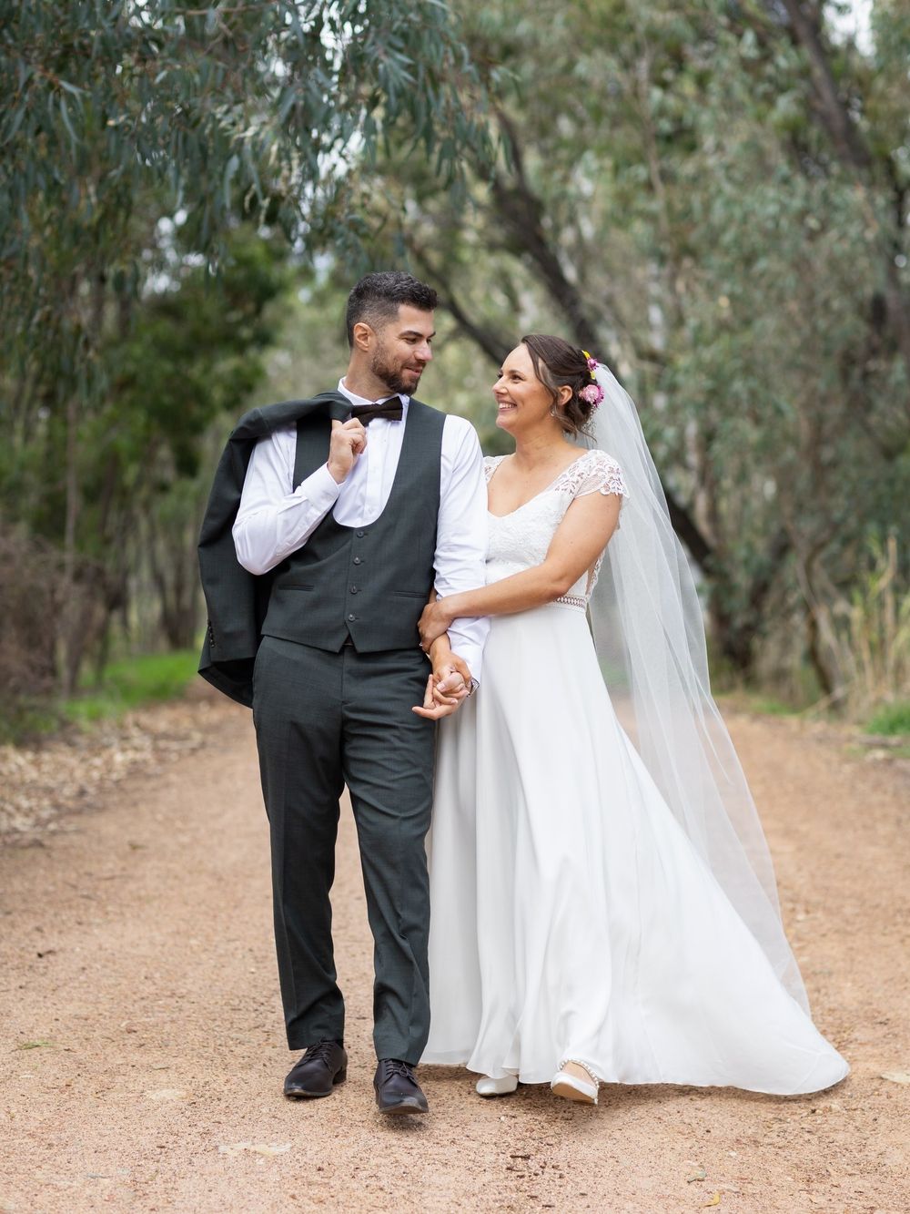 Bride and groom walking hand in hand on a dirt path surrounded by trees, smiling at each other on their wedding day.
