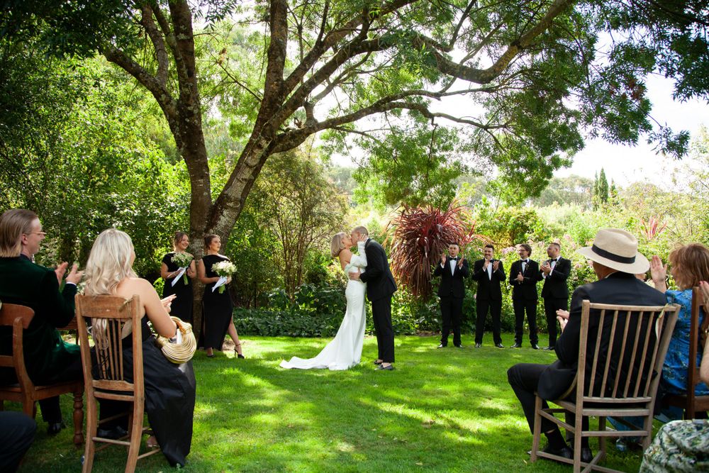 An outdoor wedding ceremony takes place under tall trees on a lush green lawn with wooden chairs for guests.