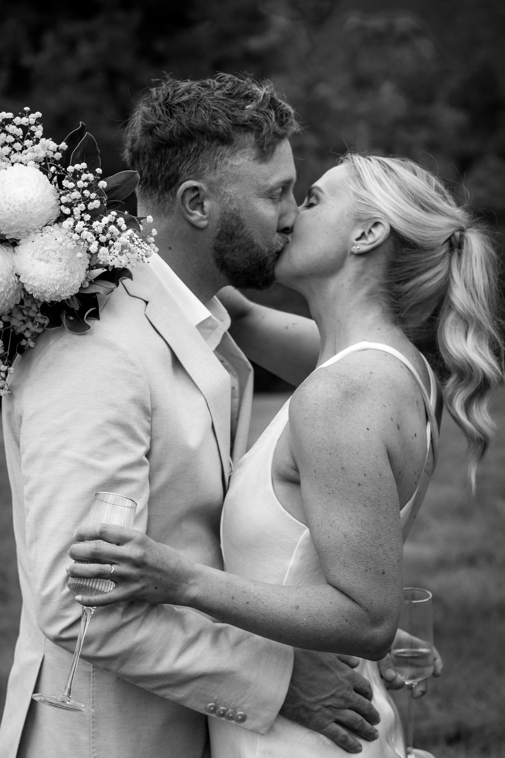 Black and white photo of a couple sharing a kiss at their wedding, bride holding a bouquet of flowers.