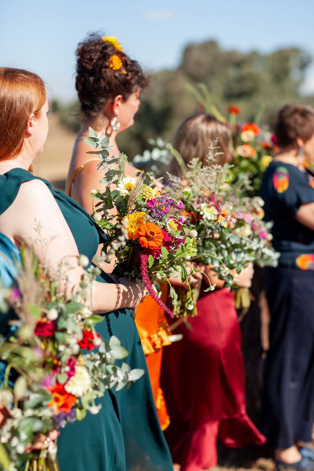 Wedding party members hold vibrant wildflower bouquets featuring orange and purple blooms while wearing jewel-toned dresses.