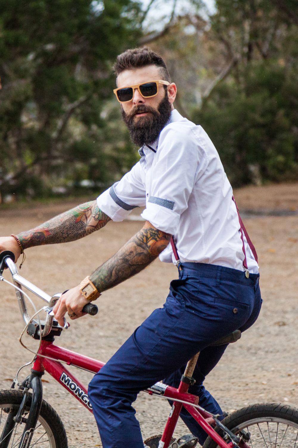 Tattooed person in white shirt and navy pants rides a red BMX bike while wearing sunglasses in an outdoor setting.