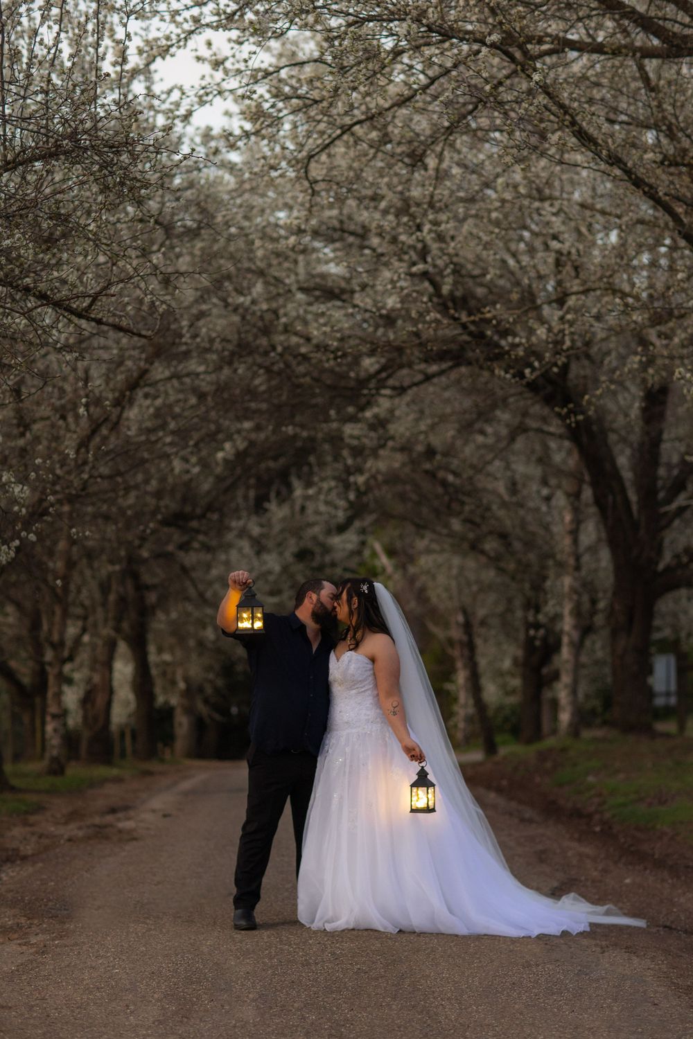 Bride and groom kissing on a tree-lined path at dusk, holding glowing lanterns in a romantic wedding portrait.