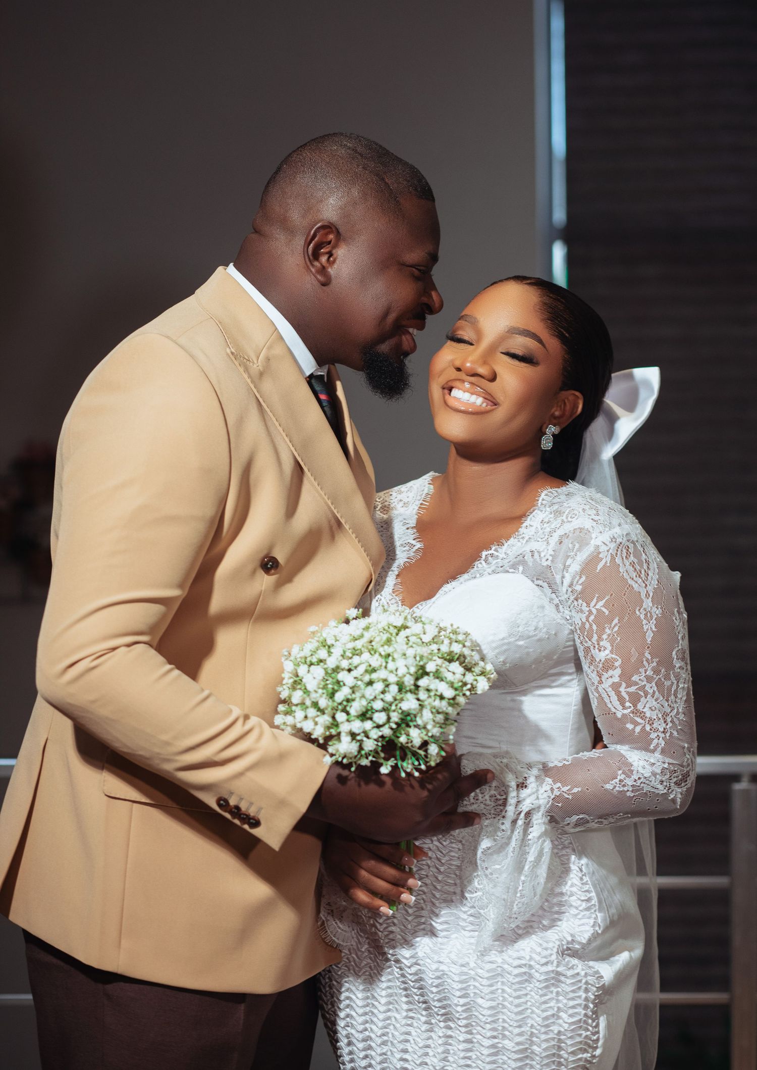A joyful wedding couple shares a tender moment while holding a baby's breath bouquet against a dark interior backdrop.