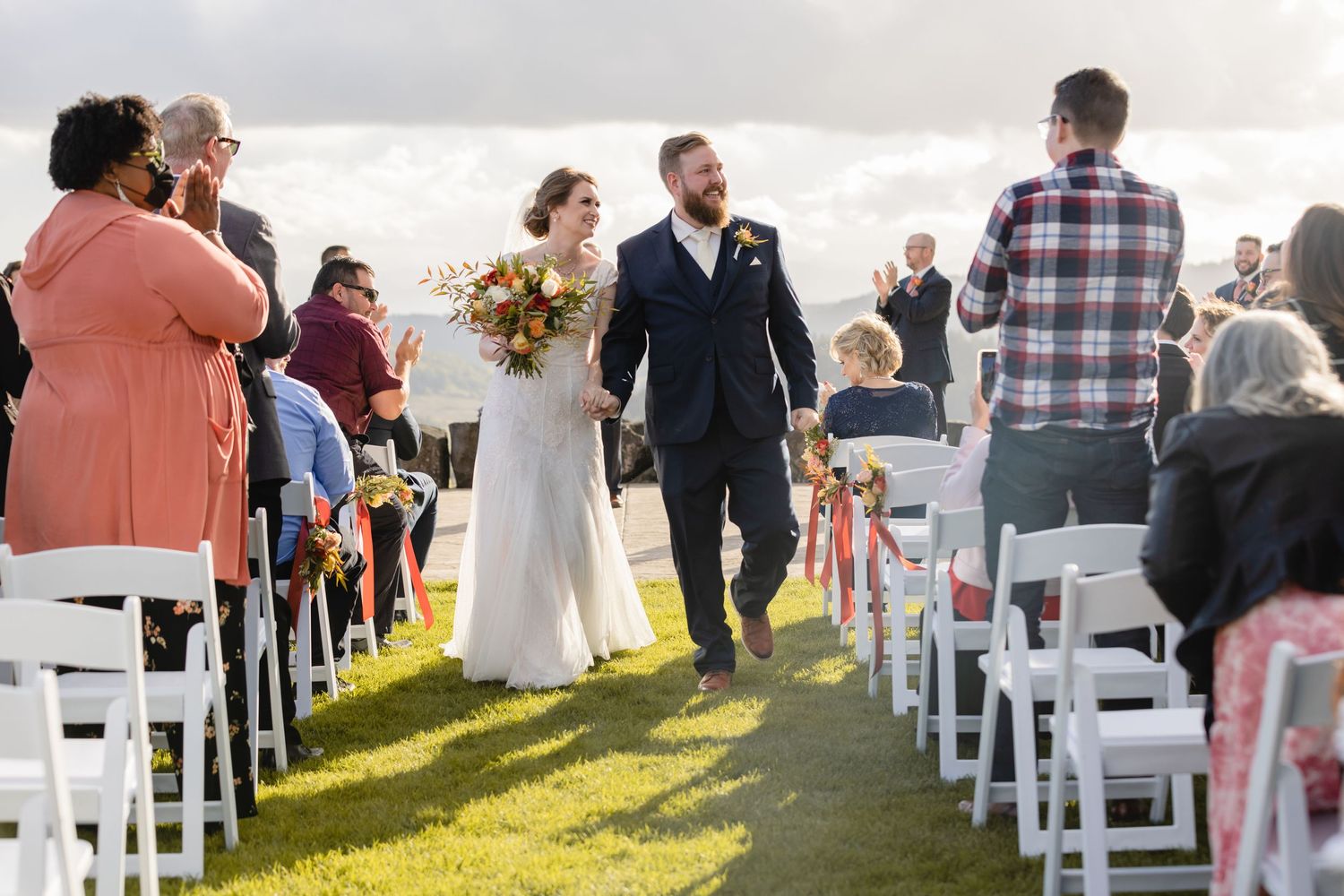 Newlyweds walk down the aisle between white chairs on a grassy hillside outdoor ceremony.