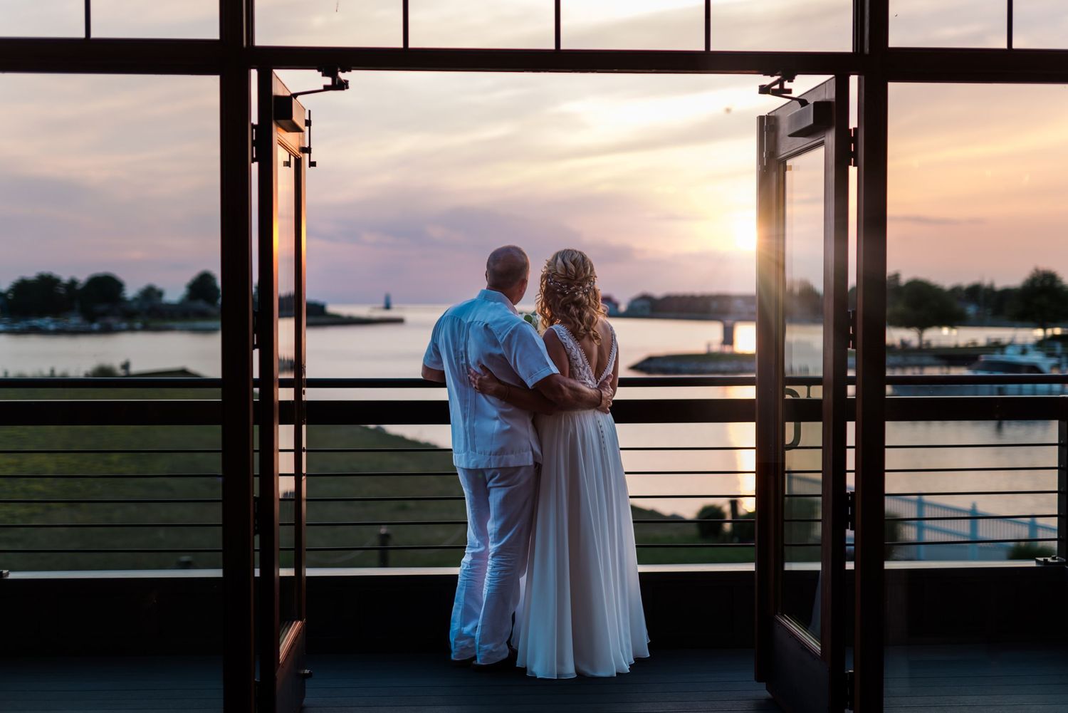A couple in matching white outfits embrace while watching the sunset from a waterfront balcony.